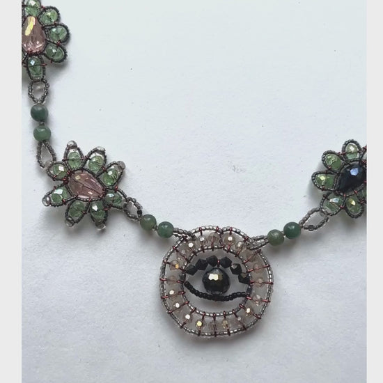 Made predominately of jade beads in varying colors with an evil eye pendant and floral shapes made of crystal beads along each side - the left side has pale pink centers while the right has black centers.