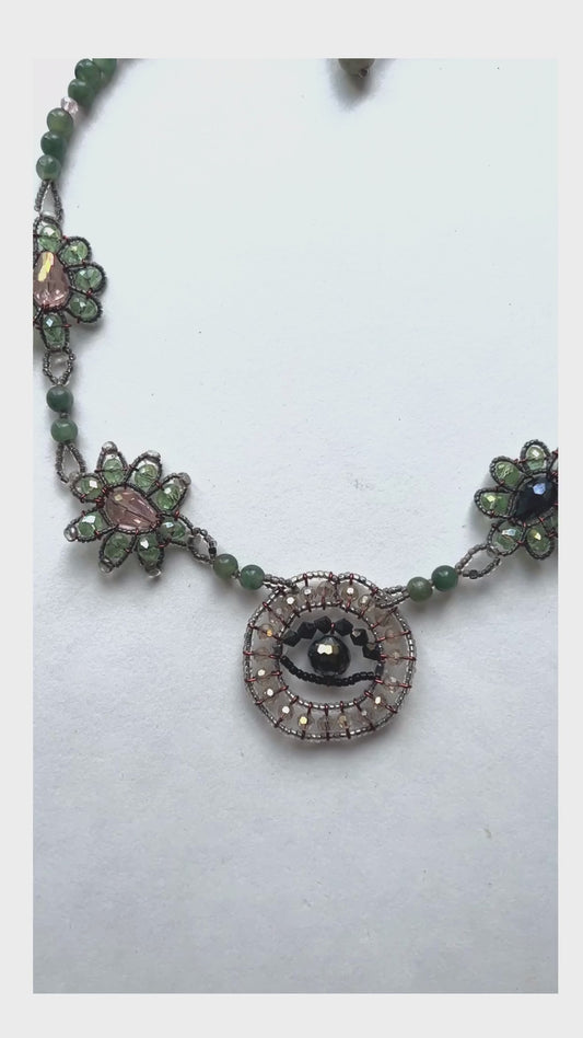 Made predominately of jade beads in varying colors with an evil eye pendant and floral shapes made of crystal beads along each side - the left side has pale pink centers while the right has black centers.